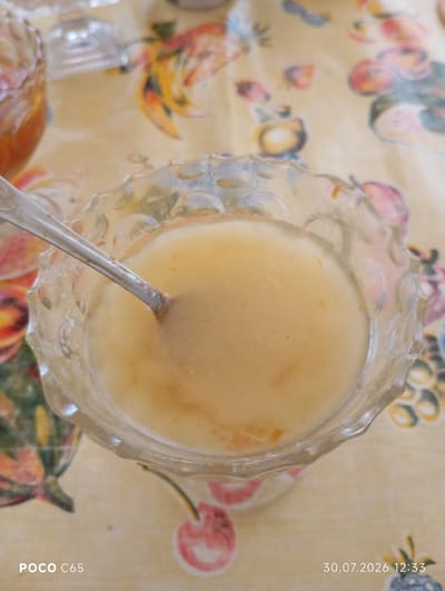 A close-up, top-down shot shows a glass bowl filled with a light orange, creamy substance, possibly a dessert or soup. A silver spoon is partially submerged in the substance. The bowl is placed on a yellow tablecloth patterned with various fruits, including cherries and what appear to be pomegranates. In the upper left corner, a portion of another glass bowl is visible, containing a darker orange substance, possibly jam or a thicker sauce. The image includes a timestamp in the bottom right corner: "30.07.2026 12:33" and a device marker in the bottom left: "POCO C65". The lighting suggests an indoor setting. No people are visible in the frame.