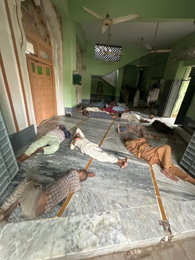 In Karachi Division, Pakistan, a group of men are lying down on a marble floor, seemingly asleep or resting. The room appears to be a hall with a high ceiling and green walls. Several ceiling fans are visible. In the background, there is a wooden door with glass panes and a window with a lattice design. The men are dressed in casual clothing, some in traditional shalwar kameez. The overall impression is a scene of repose in an interior space.