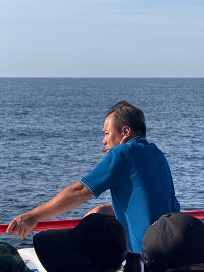 A man wearing a blue polo shirt with white trim on the collar and sleeves leans on a red railing. He is looking out at the calm blue ocean under a clear sky. His right arm is extended along the railing, and his left arm is bent at the elbow. In the foreground, two people are seated, with one wearing a black baseball cap and the other a dark hat. The scene appears to be on a boat in Dong Son, Vietnam.