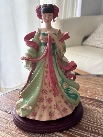 A porcelain figurine of an Asian woman stands on a wooden table. The figurine is dressed in a green robe with pink accents and floral patterns. The woman has dark hair styled in an updo with a pink accent. She is wearing dangling earrings and has her hands clasped in front of her. The figurine is on a dark red circular base. The background is blurred, showing a couch and a window with curtains. The scene is indoors, likely in a residential setting in Aventura, United States of America.