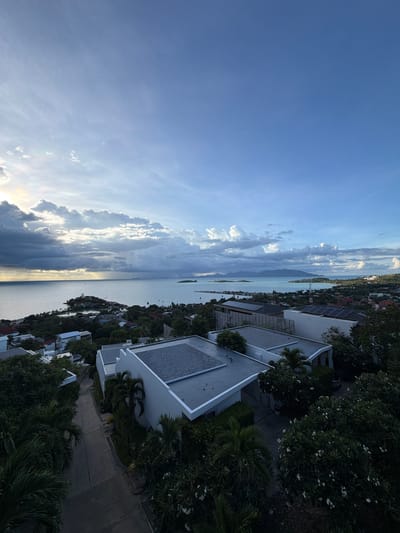 This is a high-angle, wide-shot photograph taken from a residential area in Baan Plai Laem, Thailand, overlooking the sea and distant islands. The sky dominates the upper half of the image, with a dramatic mix of dark, heavy clouds in the center and lighter, scattered clouds towards the edges, backlit by the sun which is partially obscured. The sun's rays are visible, breaking through the clouds and casting a golden glow on the water.
Below the sky is a large expanse of calm, blue-grey sea. On the horizon, several islands and landmasses are visible, including one prominent island closer to the foreground with a Ferris wheel-like structure on top. Along the coastline, houses and vegetation are densely packed, transitioning into a more developed area with modern buildings.
In the foreground and mid-ground, several contemporary houses are visible. These houses have flat roofs, some of which are covered with solar panels. The architecture is modern and clean, with white or light-colored walls. Lush green trees and palm trees surround the houses, with some flowering plants adding touches of white. A paved road winds through the residential area, bordered by houses and dense foliage. The overall impression is of a coastal residential community bathed in the soft, diffused light of either sunrise or sunset. There are no people or animals visible in the image.