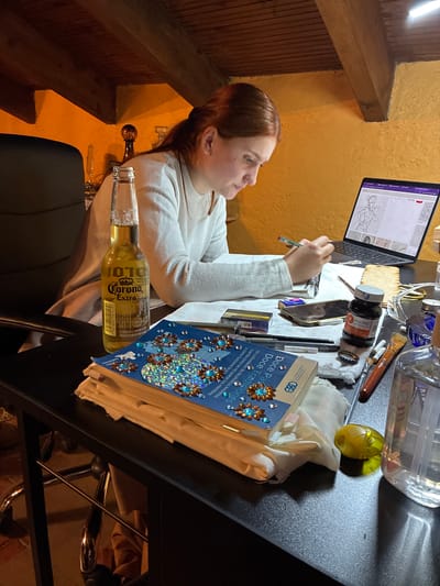 A woman sits at a desk facing a laptop with a drawing on the screen. She is wearing a white sweater and holding a pen. On the desk, there is a beer bottle, a book titled "Doce Pasos Doce Tradiciones," a bottle of ink, brushes, and other art supplies. The location appears to be indoors, possibly in a room with wooden beams and a textured wall. There is no indication of the location being Cañada de Madero, Mexico, within the image.