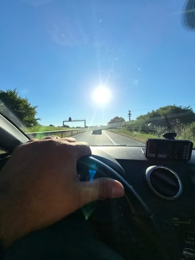 The image shows a first-person perspective of someone driving a car on a sunny day. The driver's hand is visible on the steering wheel. The road ahead is clear, with a few other cars in the distance. The sun is shining brightly in the sky, causing some lens flare. The car's dashboard is visible, with a phone mounted on it. The surrounding environment appears to be a highway with trees and vegetation on the sides. The text "VISTA 19:26 2 km 0:05 h" is visible on the car's display, suggesting navigational information or trip data. It is not possible to determine the exact location within Mondorf-les-Bains, Luxembourg from this image alone.