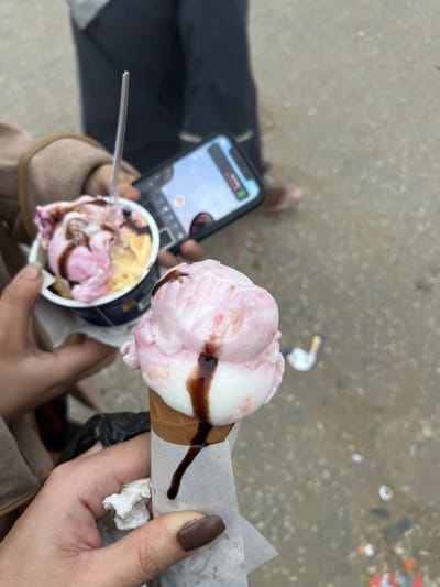 Two individuals are enjoying ice cream. One person holds a phone pointed towards a cup of ice cream with a spoon, while the other displays a pink and white cone with chocolate sauce. The nails on the cone-holding hand are painted a brown hue. Background shows pavement and scattered debris.