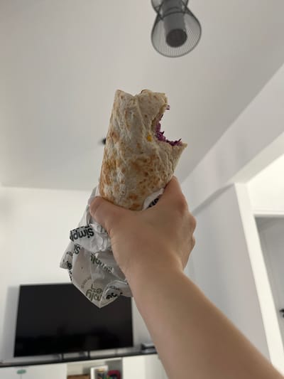 A person's hand holds a partially eaten wrap above their head. The wrap is made of a flatbread and contains visible red cabbage and corn. The wrap is partially covered in white paper packaging with green text. The background consists of a white ceiling with a modern light fixture. Below the wrap and hand, a black flat-screen television is visible on a white cabinet. The scene appears to be indoors, likely in a residential setting. The image does not contain any visual cues that would indicate a specific location within Limassol, Cyprus.