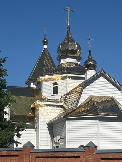 The image shows the Church of the Resurrection of Christ in Kemerovo, Russia. The church has white wooden walls and a golden roof with multiple domes, each topped with a cross. The sky is clear and blue. In the foreground, a brick wall with decorative finials can be seen. A tree with green leaves is partially visible on the left side of the image. There are no people or animals visible. The overall impression is one of a bright, sunny day and a well-maintained religious building.