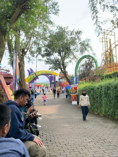The scene is at the entrance of a theme park called "Jawa Timur Park" in Batu, Indonesia (latitude: -7.885, longitude: 112.524).  The entrance is marked by a large colorful archway with the park name in large letters.  There are people walking through the entrance, some looking at their phones. A woman in a white sweatshirt and dark blue pants is walking towards the camera in the center of the path.  To the left of the path, three people are sitting on a bench. They appear to be looking at their phones and talking. The park features a variety of colorful rides and decorations, including a large green Ferris wheel in the background. The ground is paved with brick.  Trees and bushes are planted along the edges of the path. The sky is overcast.