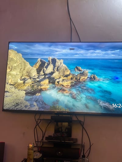 The image shows a television displaying a scenic view of rocky islands and clear turquoise water under a cloudy sky. A digital clock in the bottom right corner of the screen reads "16:24". The television is mounted on a wall above a shelf unit that holds several electronic devices and decorative items, including what appears to be a lighter with a flame and a diffuser with reeds. The environment is dimly lit, with the wall behind the television appearing to be a textured pinkish-purple. The provided image does not contain any elements that would allow for the identification of its location in Kporva, Nigeria.