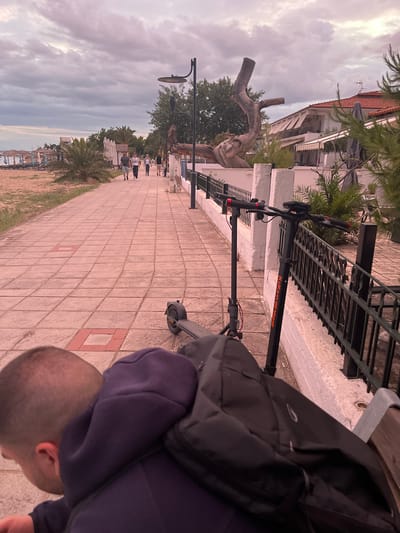 A paved promenade runs alongside a sandy beach in Gerakini, Greece. The sky is overcast with pinkish clouds. Several people are walking on the promenade, while others are seated at tables outside buildings on the right. An electric scooter is parked on the promenade, leaning against a black metal fence. A person wearing a dark purple hoodie and a backpack is seated on a bench in the foreground, facing away from the camera. The beach has lounge chairs and umbrellas.