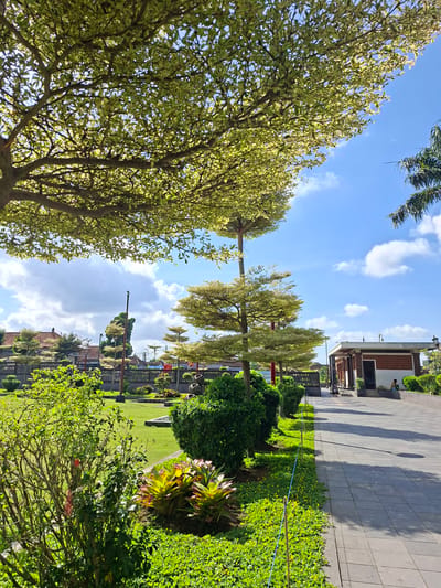The image depicts a sunny day in a park in Gianyar, Indonesia. The foreground is dominated by lush green foliage, including a large tree with a dense canopy of variegated leaves. A paved walkway lines the right side of the frame, casting long shadows from nearby trees. Beyond the walkway, a building with brick accents and a flat roof is visible. In the middle ground, a well-maintained lawn is dotted with ornamental bushes and trees. A fence separates the park from a residential area with traditional Balinese architecture in the background. Two individuals are present; one is seated on a low wall near the building, and another appears to be walking in the distance. The sky is a vibrant blue with scattered white clouds.