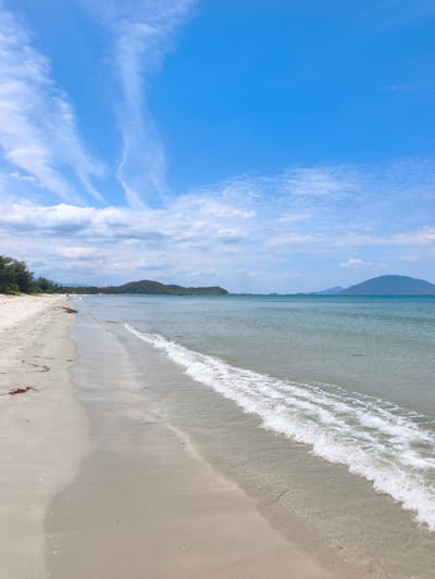 A wide, sandy beach stretches along the coast, meeting a calm, blue sea. Gentle waves with white foam lap at the shore. In the distance, a line of lush green hills and mountains borders the horizon. The sky is a bright blue, dotted with wispy white clouds. A few scattered pieces of dark seaweed lie on the wet sand near the water's edge. The scene is serene and evokes a sense of natural beauty. The location is identified as Dong Ninh Hoa, Vietnam.