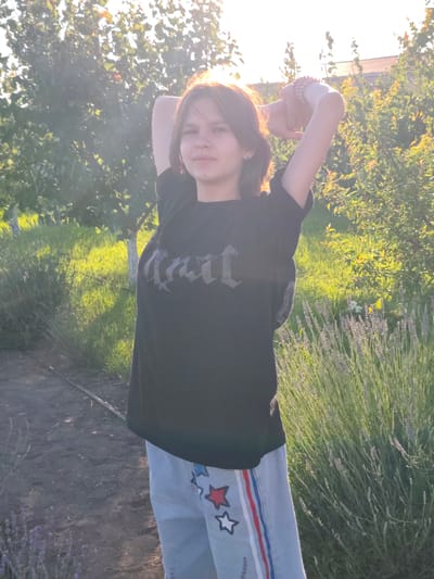 A young person with medium-length brown hair stands in an outdoor setting. They are wearing a black t-shirt with a gothic-style text graphic and light-colored pants adorned with red and white stars and stripes. Their arms are raised above their head, with hands clasped behind their neck.
The scene appears to be a garden or yard, with trees and lush green foliage visible in the background. A patch of lavender is in bloom to the right of the subject. The lighting suggests it is late afternoon or early evening, with the sun casting a warm glow and creating some lens flare. The ground in front of the subject is a dirt path. The overall mood seems relaxed and peaceful. No specific event is discernable, but the posture might indicate stretching or posing. The text on the t-shirt is in a stylized font and appears to be a word or name in a gothic script, but it is not clearly legible.