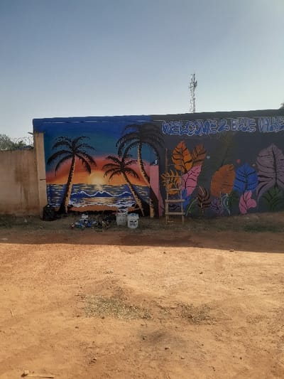 This image depicts an outdoor daytime scene in Kirana, Nigeria, centered on a long, concrete wall serving as a mural canvas. The ground in the foreground and midground consists of reddish-brown dirt with sparse, dry vegetation.
The wall, approximately 2 meters in height, is divided into two distinct mural sections. The left section portrays a tropical sunset scene with two prominent black silhouettes of palm trees flanking a sun setting or rising over a body of water with white-capped waves. The sky transitions from orange and yellow hues near the sun to a deep blue at the top. Below this mural, on the dirt ground, a black plastic bag, several small pieces of refuse, and two white plastic paint buckets are visible.
The right section of the wall features a dark grey background with stylized text in blue and white, reading "WELCOME 2 BLUE VALLE". Below the text, several large, stylized leaves are painted in shades of orange, brown, purple, pink, blue, and light green. A small, three-step wooden ladder leans against the wall beneath this mural.
In the far background, against a clear, pale blue sky, the upper structure of a metallic lattice communication tower is visible above the wall. To the far left, behind the wall, a light-colored building structure topped with barbed wire is partially visible. No individuals are present in the frame.