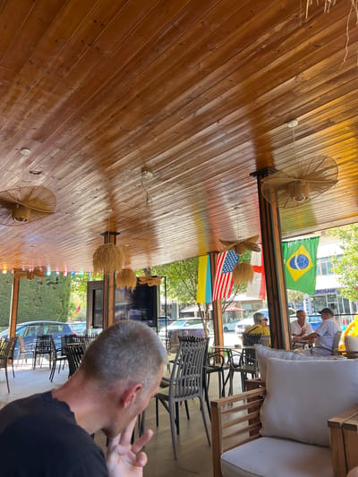 The image depicts an outdoor seating area of a cafe or restaurant with a wooden ceiling. Several tables and chairs are visible, with some patrons seated at one table in the background. Flags of various countries, including the United States and Brazil, are displayed. A man in the foreground is looking down and gesturing with his hand. The environment appears to be a city street with cars and buildings visible in the background. The overall impression is a casual, public gathering space. The location is identified as Bashkia Durrës, Albania.