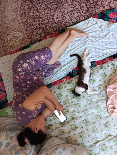 A woman is lying on a bed with her legs up, leaning against a wall or headboard. She is wearing a purple floral dress and holding a smartphone. A black and white cat is sleeping on the bed next to her. The room is decorated with floral-patterned fabrics. The image appears to be taken indoors during the day. The woman's posture suggests relaxation, and the presence of the cat adds a sense of domesticity. The location is specified as Nukus, Uzbekistan. The smartphone screen is visible but displays only a blank white interface, so no text can be read from it.