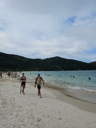 This image depicts a vibrant beach scene at Prainhas do Pontal do Atalaia in Arraial do Cabo, Brazil. The sky is partially cloudy with visible blue patches. In the foreground, two individuals walk barefoot along the pale sand from left to right. A woman, wearing a black bikini top and black shorts, holds a green object resembling a rolled beach mat or umbrella pole in her left hand and a translucent cup in her right. To her right, a man with a blue baseball cap and a light-colored t-shirt wears a dark backpack and carries a dark pouch on his side. He wears light-colored flip-flops.
The expansive beach extends into the midground, populated by numerous other individuals either walking along the shoreline or congregating further up the sand, near what appear to be vendors or shaded areas. The ocean water, visible to the right of the beach, is a clear turquoise color with gentle waves breaking near the shore. Numerous people are seen swimming or wading in the water. In the distance on the water, at least two white motorboats are visible. Lush, green, tree-covered hills form the backdrop, sloping down towards the ocean on both sides of the bay, framing the entire scene.