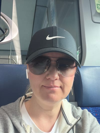 A woman wearing a black Nike baseball cap and sunglasses is sitting on a blue seat in what appears to be a train. She has white earbuds in her ears and is wearing a grey hooded jacket over a white t-shirt. The background shows the interior of a train car, with blue seats and windows. The lighting suggests it is daytime, with sunlight casting shadows on the windows. The woman is looking towards the camera and smiling slightly.