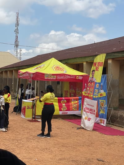 This is a vertical, eye-level, outdoor shot of a promotional event taking place in a dusty, open area, possibly a market or community space. The time of day appears to be midday, with bright sunlight casting shadows. A large, yellow and red tent serves as the focal point, adorned with vibrant banners advertising "Chupa Chups" candy. The banners feature images of candy packs and people engaged in playful activities, with text in various colors, including English slogans like "Stretch. Twist. Toss.", "Your Fun.", "Roll on", "Toss on", and "Available in any shop near you!".
Several people are visible, mostly young adults, dressed casually. One woman, wearing a yellow Chupa Chups branded shirt and black pants, is actively engaged, holding a phone as if taking a picture or video. Others, some in similar branded shirts, are positioned behind a counter or table, suggesting they are part of the promotion. The overall atmosphere seems energetic and promotional, with a focus on engaging the public.
In the background, a building with a corrugated iron roof and an older facade is visible. Beyond that, a telecommunications tower stands tall against a partly cloudy sky. The ground is a reddish-brown dirt, typical of arid or semi-arid regions. The presence of the television tower hints at a connection to broadcasting or media events, and the mention of Nigeria in the location context further grounds the scene. The scene conveys a sense of local community engagement with a global brand.
