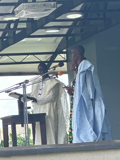 In Itoga, Nigeria, three people stand on a raised platform, suggesting an outdoor or semi-outdoor event. The central figure, facing left, wears a cream-colored garment with intricate embroidery on the chest. To their right, a person in a light blue, patterned robe, also facing left, appears to be listening or speaking. Further to the right, a third individual, partially obscured, faces forward. Microphones are positioned in front of the central figure, and a wooden lectern is visible to their left, indicating a speech or sermon. Overhead, a white canopy is supported by metal beams, and circular light fixtures are embedded in the structure, providing illumination. The background consists of a muted blue wall and the suggestion of trees and a building beyond the platform.