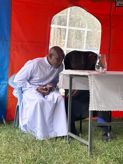 This image depicts an outdoor gathering under a tent in Kiganjo, Kenya, illuminated by bright daylight. The interior of the tent features red and blue fabric walls, with a clear plastic window offering a view of a grassy outdoor area and distant structures.

Two men are visible. In the foreground, a dark-skinned, bald man is seated on a light blue plastic chair, wearing a light-colored, long-sleeved robe or tunic, likely white or light blue. His hands are clasped in front of him, holding what appears to be a pen, and he is looking upwards and to the right, seemingly attentive to something outside the frame. Behind a table draped with a white, decorative tablecloth, another dark-skinned man is partially visible, wearing a dark top.

On the table, a clear plastic bottle containing a liquid, possibly a dispenser for water or hand sanitizer, is present. The metal frame of the table is also visible. The ground outside the tent is covered in vibrant green grass, indicating a sunny day.

The setting, including the tent, the formal attire of the man in the foreground, and the visible text "Methodist" on the red tent fabric in the upper left corner, strongly suggests a religious or community event, likely associated with the Methodist church. The atmosphere appears calm and observant. Additional visible text on the tent includes "0721".