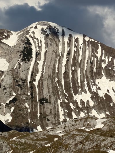 The image depicts a steep, rocky mountain face with visible horizontal striations and patches of snow. The mountain appears to be partially shadowed, with darker rock formations. A portion of the mountain is covered in moss and vegetation. The sky is visible in the background, suggesting a cloudy or overcast day. It does not show identifiable characteristics that would indicate the city of Boričje, Montenegro.