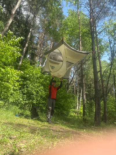 The image shows a man standing in a forest in Папярнянскі сельскі Савет, Belarus, holding up a tent. The tent appears to be hanging from a tree branch. The forest has many green trees and bushes. The sky is visible through the trees, with some blue patches. The man is wearing a red shirt, grey pants, and black shoes. He seems to be in the process of setting up or adjusting the tent.