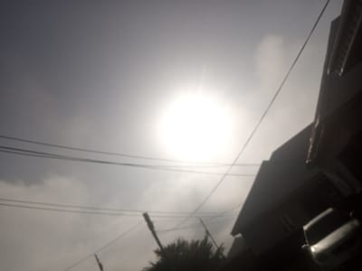 The image depicts a hazy outdoor scene, likely in Katon Rikkos, Nigeria. The most prominent element is the bright, washed-out sun in the upper center of the frame, suggesting it is daytime, possibly midday or late afternoon, with the sun high in the sky. The sun's glare creates a halo effect and obscures details in the sky.
Overhead, multiple thin wires crisscross the frame, indicating the presence of utility lines, likely for electricity or telecommunications. Below the wires, to the right, is a dark, angled structure that appears to be the roof and upper part of a building, possibly a house or commercial establishment. The building's roof has a distinct, stepped or layered design, and a small section of what looks like a window or opening is visible. Further to the right and lower, a portion of a vehicle is visible. It's a light-colored car, possibly silver or white, with reflective surfaces, including what appears to be a side mirror.
In the lower part of the image, a dark, bushy mass of trees is partially visible. The overall atmosphere is somewhat muted and hazy, suggesting a cloudy or dusty sky, or possibly the atmospheric conditions common in certain seasons in Nigeria. The quality of the image is somewhat degraded, with a lack of sharp focus and a prevalence of light and shadow. No people are clearly visible. The scene conveys a sense of a typical day in an urban or semi-urban area, with infrastructure and daily life elements present, albeit blurred by atmospheric conditions and the strong sunlight. There is no visible text in the image.