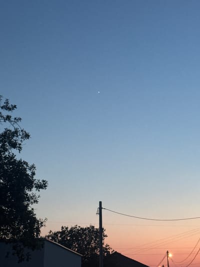 This image captures a twilight sky over Sustaš, Montenegro. The sky transitions from a deep blue at the top to a soft orange and pink gradient near the horizon. A single bright star or planet is visible in the upper portion of the sky. Silhouetted against the sky are the branches of a tree on the left, a utility pole with wires in the middle, and the roofline of a building and more trees on the right. The overall scene suggests the end of the day.