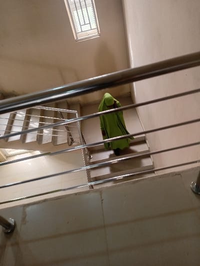 The image depicts an individual descending an interior stairwell within a building in Kwazailewa, Nigeria. The subject is viewed from an elevated perspective, positioned with their back to the camera, wearing a vibrant green full-body garment that covers their head and form. Dark footwear is visible beneath the garment. The stairwell features light-colored tiled steps and walls. A modern metal railing system, composed of multiple horizontal bars, traverses the mid-ground. A rectangular window with a visible grid pattern is situated high on a wall, providing ambient illumination. A light-colored tiled wall section forms the immediate foreground, anchoring the railing.
