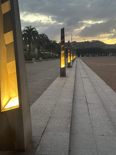 A wide, grey stone promenade, stretching diagonally from the bottom-left to the middle-right of the frame, dominates the scene. The promenade is composed of large, rectangular stone slabs with visible seams. Along its right edge, a series of modern, rectangular light fixtures are integrated into a low stone wall or structure, casting a warm, bright yellow glow onto the path and creating strong linear shadows.
To the left of the promenade, a vast, flat expanse of sandy ground extends into the distance, possibly indicating a beach or a large open area. In the mid-ground, further to the left, several dark green palm trees are visible, silhouetted against the sky.
The background on the far right features a distant hill or mountain, covered in vegetation and dotted with scattered buildings, suggesting a coastal town or city. The sky above is partially cloudy, with lighter, warm-toned patches breaking through darker clouds, characteristic of either dawn or dusk. The artificial lights are prominently lit, reinforcing the low-light conditions. The overall mood is calm and urban, with no people or specific activities depicted. No text is visible in the image. The setting is a paved waterfront area in Gijón, Spain.