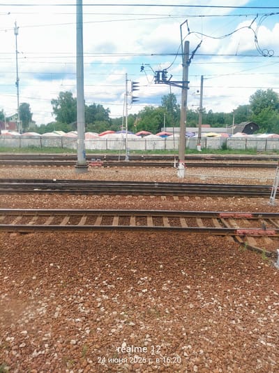 The image shows a railway track with multiple sets of rails on a bed of gravel. Overhead, numerous power lines and support structures for the electric railway are visible against a blue sky with scattered clouds. In the background, a line of white tents or market stalls is set up behind a metal fence. Beyond the stalls, trees and some buildings are present, including a dark, angular structure that appears to be a small barn or shed. The scene suggests an outdoor setting with evidence of both infrastructure for transportation and a commercial or recreational area.

The time of day is indicated by the watermark "16:20" which, along with the bright sky and shadows, suggests it is late afternoon. The weather appears to be fair with a partly cloudy sky. The presence of the train tracks and overhead electrical lines indicates an active railway line, possibly at a station or near a depot. The tents in the background could signify a market or an event taking place. No people are clearly visible in the image.

The visible text includes "realme 12" and "24 июня 2026 г в 16:20" which is a date and time stamp in Russian, indicating "June 24, 2026 at 16:20". The context provided states the location as Monino, Russia.