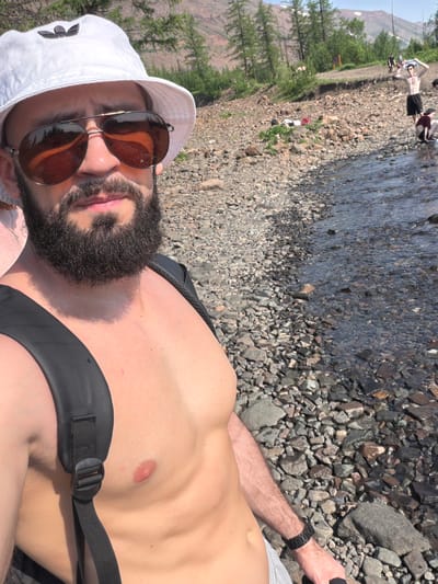 A man with a dark beard and sunglasses, wearing a white bucket hat and a black backpack, is standing on a rocky shore next to a body of water. In the background, several other people are visible, some are walking on the shore and some are sitting down. Trees and mountains are in the distance. The scene appears to be a natural landscape, possibly a riverbank or lake shore in a mountainous region. Given the context of Talnakh, Russia, it is likely a location near the city, which is known for its subarctic climate and proximity to natural wilderness.