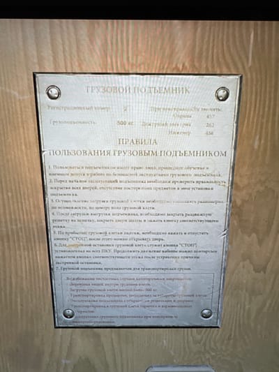 A metallic sign, likely brushed aluminum or stainless steel, is prominently displayed on a wooden wall with a visible grain pattern. The sign is affixed with four screws or rivets, one in each corner, and shows minor signs of wear such as scratches and smudges. The lighting suggests an indoor setting.

The text on the sign is in Russian and details information and rules for using a freight lift. The main title is "ГРУЗОВОЙ ПОДЪЕМНИК" (FREIGHT LIFT). Key specifications include a registration number "2" and a load capacity of "500 кг" (500 kg). Other numbers like "263" and "456" are also listed, though their specific meaning is unclear from the image alone.

Under the heading "ПРАВИЛА ПОЛЬЗОВАНИЯ ГРУЗОВЫМ ПОДЪЕМНИКОМ" (RULES FOR USING THE FREIGHT LIFT), a series of instructions are provided. These rules cover:
1.  Operation by trained and authorized personnel only.
2.  Pre-operation checks, including door closure and absence of foreign objects.
3.  Proper loading of cargo, ideally centered on the lift floor.
4.  Correct procedure for closing gates and doors after loading/unloading.
5.  Using the "СТОП" (STOP) button upon arrival at a floor before opening doors.
6.  The function of the "СТОП" button as an emergency stop on all control panels and how to resume operation.
7.  The primary purpose of the lift for transporting goods, explicitly prohibiting the transport of people, loads exceeding 500 kg, items protruding beyond the lift's dimensions, and corrosive or explosive substances.