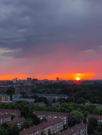 This image captures a vibrant sunset over the city of Rijswijk, Netherlands. The sky is a dramatic canvas of deep grays and purples at the top, transitioning to a brilliant gradient of pinks and fiery oranges near the horizon. The sun, a bright orange orb, is prominently visible, just beginning to dip below the skyline.
The cityscape below is a mixture of residential buildings and green spaces. In the foreground, several multi-story apartment buildings with reddish-brown roofs are visible. These buildings feature numerous windows and balconies. Further in the background, more apartment blocks and some taller structures, possibly office buildings or hotels, punctuate the landscape. A distinctive, modern white building with a triangular roof is also visible on the left side of the city. The horizon is lined with trees, forming a dense, dark green band that separates the sky from the urban environment. A construction crane stands tall among the buildings, suggesting ongoing development in the city. The overall scene is one of an urban twilight, bathed in the warm, dramatic light of the setting sun. No people are visible in the image.