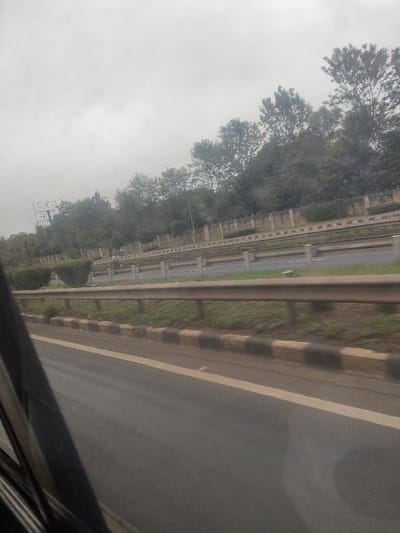 The image depicts an outdoor road scene in Gatongora ward, Kenya, under an overcast sky. The perspective suggests it was taken from a moving vehicle.
In the immediate foreground, a segment of an asphalt road is visible, featuring a solid white lane marking on its left edge. This road section is bordered by a curb painted with alternating black and pale yellow-beige stripes.
Beyond this, a horizontal, multi-sectional galvanized metal guardrail spans the width of the frame. Behind this guardrail, a grassy median contains several low, trimmed green bushes.
Further into the midground, an asphalt highway with multiple lanes is visible, separated by another median barrier. This barrier consists of multiple individual posts supporting horizontal metal rails. On this secondary median, a single individual is discernible, wearing high-visibility clothing, specifically a yellow or lime green vest. The person appears to be standing or slowly walking on the median.
The background features a substantial wall, possibly constructed from concrete or stone blocks, which runs parallel to the road and appears to have security enhancements, such as a top layer of barbed wire or similar deterrents. Behind this wall, a dense line of tall, mature green trees forms a forested backdrop. To the far left in the background, a large, empty billboard structure with a blue metal frame is partially visible above the tree line. A utility pole or street light extends upwards from the foliage on the right side of the frame.