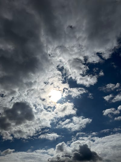 The image shows a dramatic sky over Męcina, Poland, with a mix of dark, heavy clouds and bright, fluffy cumulus clouds. The sun is partially obscured by the clouds, creating a bright glow and casting shafts of light. The sky is a deep blue, visible through the gaps in the clouds. The overall impression is of a dynamic and somewhat moody sky, suggesting changing weather conditions. No people, objects, or specific landmarks of Męcina are visible in the frame, as the image is solely focused on the sky.