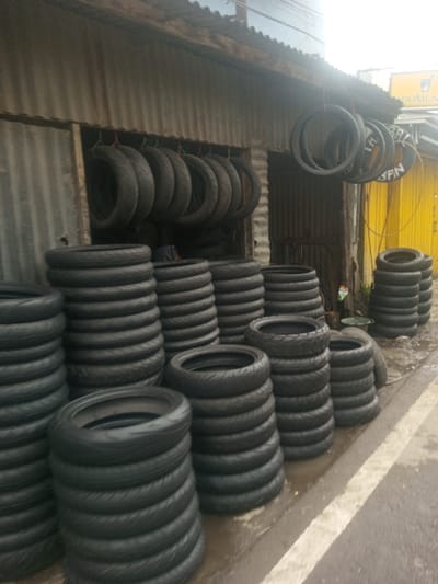 This image depicts a tire shop located in Sarwahi Mal., India. The scene is dominated by numerous black vehicle tires, primarily consistent with motorcycle or scooter types. These tires are organized into multiple vertical stacks, approximately 10 to 15 tires high, positioned on a wet, dark-colored ground surface that exhibits a partial white road marking. Additional tires, numbering approximately 12 to 15, are suspended from hooks and ropes beneath a corrugated metal roof overhang that extends along the shop's facade. The shop building features walls constructed from corrugated metal panels and an open entrance on the left, revealing more tire inventory inside. To the right of the shop, an adjacent structure presents a prominent yellow corrugated metal wall, a dark doorway, and an indistinct sign partially visible above it. A green basin is situated on the ground near the base of the yellow wall. No individuals are present in the depicted scene.