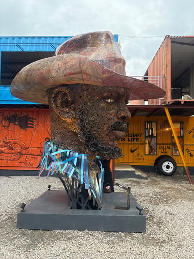 This image depicts a large outdoor sculpture of a man's head and shoulders, crafted from metal, in Karum, Nigeria. The sculpture is positioned in front of several shipping containers. A blue shipping container is visible to the left, partially obscured by the sculpture. To the right of the sculpture, an orange shipping container with a mural is on the left, and a yellow food truck with text and graphics is on the right. The sculpture features a weathered hat, detailed facial features constructed from coiled metal, and a collar made of colorful, ribbon-like metal strips. The ground is covered with gravel. Two small spotlights are visible at the base of the sculpture's platform. The sky is overcast.