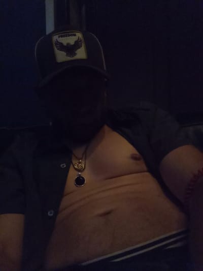 An adult individual, appearing male, is depicted from the chest up in a dimly lit interior setting. The person is wearing a dark baseball cap featuring a light-colored square patch with a dark eagle silhouette and the word "FREEDOM" above it; the cap casts a deep shadow, obscuring the face. A dark short-sleeved button-up shirt is worn unbuttoned, revealing the individual's chest and abdomen. Two necklaces are visible: one with a gold-toned, intricately designed circular pendant, and another with a dark, possibly black, circular stone pendant. A red beaded bracelet is present on the right wrist. The waistband of dark underwear with a prominent horizontal white stripe is visible below the exposed abdomen. The individual is reclined or seated against a dark, featureless background, possibly an upholstered surface. The specific location within Seattle, United States, is not discernible from the visual evidence provided in the image.