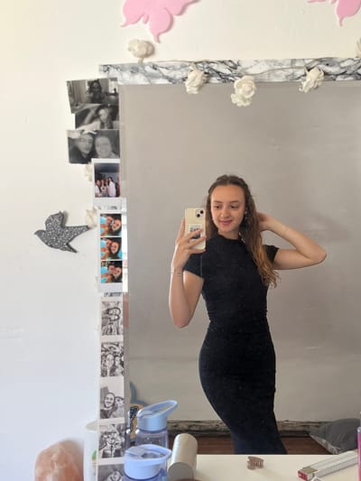 A young woman is taking a selfie in a mirror. She is wearing a black, form-fitting dress. The mirror is decorated with marble-patterned trim, artificial flowers, black and white photos, a sequined bird, and pink butterfly decorations. In the reflection, part of a desk or table is visible with items such as water bottles, a roll of tape, a hair clip, and a measuring tape. The location is implied to be in Saarbrücken, Germany, based on the prompt.