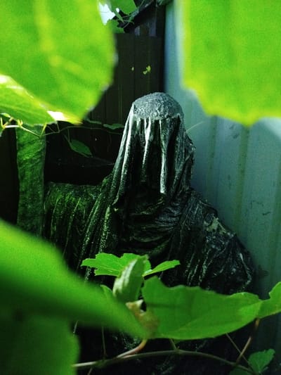 The image depicts a cloaked, seated figure, possibly a statue or effigy, partially obscured by vibrant green foliage. The figure is dark and textured, with a draped hood covering its head and shoulders, suggesting a somber or mysterious presence. The surrounding environment appears to be an outdoor setting, possibly a garden or overgrown area, with the lush green leaves creating a natural frame for the central subject. The lighting is dim, with a greenish hue, and the background consists of a textured wall and dark wooden structures. There is no identifiable indication of the specific location in Taganrog, Russia.