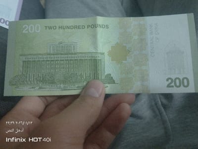The image shows a 200 Syrian Pound banknote. The bill is light green and features "200 TWO HUNDRED POUNDS" printed on it. A building is depicted on the left side of the note, with an ornate geometric pattern and "CENTRAL BANK OF SYRIA" printed on the right. The number "200" is also displayed on the lower right corner. The text "۲۲:۳۹ ۲۰۲۶/۱۰/۱۳ أدخل النص Infinix HOT 40i" appears at the bottom of the image. A hand is holding the bill.