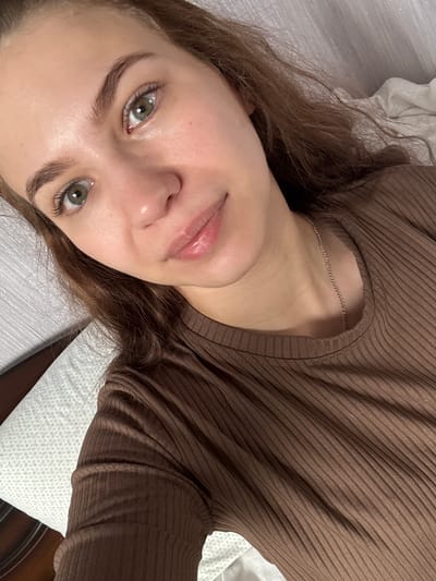 A young woman is lying on a bed or similar surface. She is wearing a brown ribbed shirt and a delicate silver necklace. Her hair is light brown and slightly wavy. Her eyes are green. The background includes a white patterned pillow and a hint of dark brown furniture. The photograph does not offer any context to identify a location in Nizhnyaya Gar', Russia.
