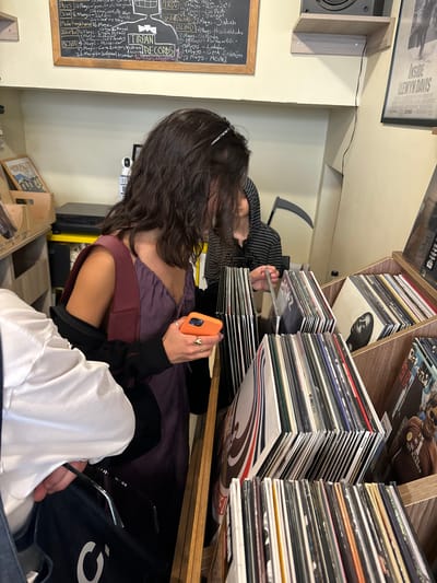 A medium-shot angled downward captures a moment in a record store in Kadıköy, Turkey. A woman with long dark hair, wearing a purple dress with burgundy straps, is holding an orange smartphone and looking at a stack of vinyl records. A second person is visible across the records from her. A chalkboard on the left lists Turkish bands performing in May, specifically mentioning Tirpan Records and Z-AP, and includes information such as days and venue for each concert. The chalkboard reads as a list of Turkish artists: "TIRPAN", "Zorlu", and "BOVA". The venue, "TKSV" also listed. Other details listed on the chalkboard include artist names like Gaye Akyol and bands like Miskinler. A poster of the film "Inside Llewyn Davis" hangs above the speaker. The environment is dimly lit, suggesting an intimate record store setting. The area is filled with shelves of vinyl records. The image captures the woman's engagement with the records.