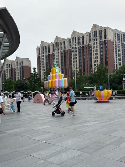 In Haidian District, China, a public square features a vibrant, oversized teacup sculpture as a focal point. The teacup, adorned with colorful stripes of yellow, pink, and orange, sits on a saucer and appears to be a popular spot for photos, with people gathered around it. Adjacent to the teacup is a multi-tiered, cylindrical sculpture resembling a colorful cake, decorated with pastel-colored, balloon-like elements and the word "DREAM" in neon lights. Several individuals, including adults and children, are present in the square, some pushing strollers, others walking, and a few children playing. The square's ground is paved with large, grey stone tiles. In the background, numerous tall, modern apartment buildings with red brick accents and numerous windows dominate the skyline, suggesting a densely populated urban environment. A large, curved, modern structure with a grey, metallic roof is partially visible on the left side of the image, possibly part of a commercial building or public facility. The sky is overcast, contributing to a diffused light across the scene.