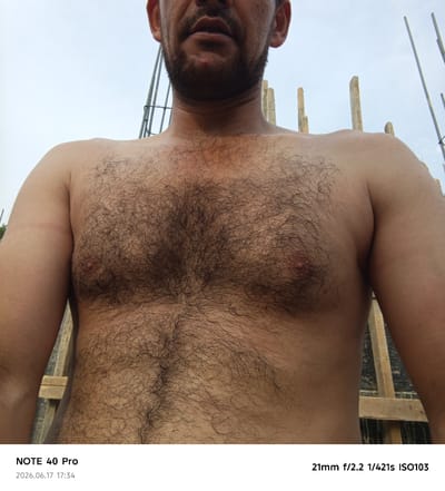 The image is a close-up, waist-up shot of a shirtless man with dark, thick chest hair. He has a fair complexion and appears to be outdoors, with a bright, overcast sky as the background. Construction materials, including wooden beams and metal rebar, are visible behind him. The man's face is not fully visible, but his mouth and chin area are in the frame, showing some stubble. The timestamp "2026.06.17 17:34" and camera information "NOTE 40 Pro 21mm f/2.2 1/421s ISO103" are overlaid on the bottom of the image. The location of Kyzyl-Yulduz, Uzbekistan is stated in the prompt but not visually depicted within the image.