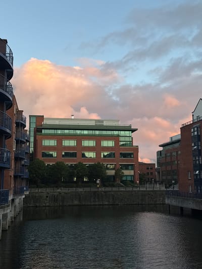 The image depicts an urban waterfront scene at dusk, likely in Dublin, Ireland, given the architectural style and presence of a canal. The sky is a soft blue with large, fluffy clouds illuminated by the setting sun, casting a warm, peachy glow on them.
Dominating the frame are several modern brick and glass buildings lining the waterway. These buildings appear to be multi-story office or residential structures with large windows reflecting the sky and surrounding environment. Some have green tinted glass. Trees and greenery are visible between and around the buildings, adding a touch of nature to the urban landscape.
In the foreground, a row of brick buildings with dark, arched structures topped with metallic cages suggests an older architectural style or perhaps a functional element of the waterfront, possibly related to flood control or infrastructure. These structures have a utilitarian, industrial aesthetic.
The water in the canal is dark and textured, reflecting the sky and buildings. Ripples on the surface suggest a slight breeze. The overall mood is tranquil and serene, evoking a sense of the day winding down. The lighting suggests it is late afternoon or early evening. There is no visible text in the image.