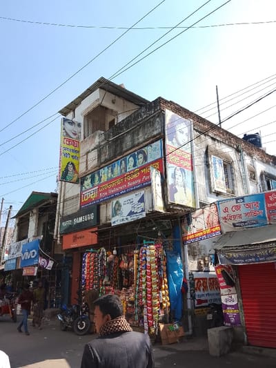 An urban street scene showcases a multi-story building with vibrant commercial signage. Businesses advertised include "Keya Makeovers Beauty Parlour" and "Samsung." Below, a shop displays rows of packaged snacks. People walk along the street, and a parked motorcycle is visible. The sky is clear and blue, and overhead power lines crisscross the scene. The buildings show signs of age, with some areas appearing weathered.