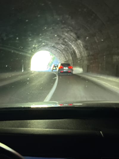 The image shows the interior of a vehicle driving through a tunnel. The tunnel is arched and appears to be constructed of rough stone. There is bright light at the end of the tunnel, suggesting an exit. Two cars are visible ahead, one dark-colored and the other a lighter shade, both appearing to be moving through the tunnel. The interior of the vehicle in the foreground shows a dashboard and windshield, with dust or particles visible on the glass. The overall impression is of a journey through a shaded, somewhat dimly lit passageway leading towards daylight. This scene could plausibly be part of the road network in or around Mišići, Montenegro, given the mountainous terrain and the presence of tunnels in the region.