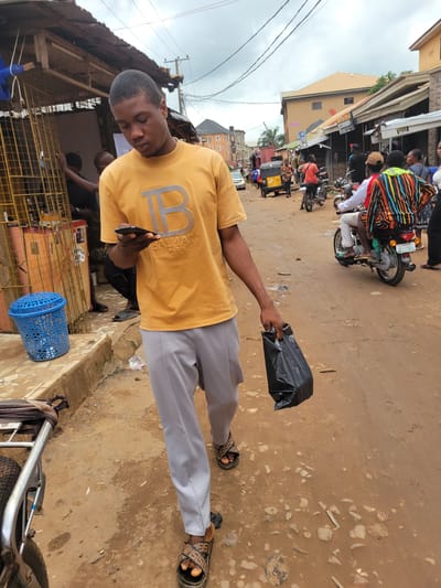 A person in a yellow shirt and gray pants stands on a dirt road in what appears to be a market or busy street in Egbagu, Nigeria. The person is holding a black plastic bag in their right hand and a smartphone in their left hand. The yellow shirt has a large silver "B" logo with "BALMAIN PARIS" written below it. The person is wearing sandals.
In the background, there are other people, some on motorcycles, and buildings that suggest a commercial area. A motorcycle with a colorful striped fabric draped over its rear is visible, as is a yellow tricycle or "Keke Napep." The ground is dusty and uneven, with some scattered debris. A blue plastic basket is also visible on the ground to the right of the person.
The lighting suggests it is daytime, possibly overcast given the sky's appearance. The overall scene depicts a slice of daily life in a Nigerian market setting. There is no obvious emotional expression on the person's face, but they appear to be looking down at their phone.